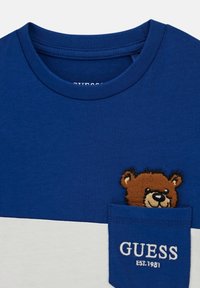 Mėlyna medvilninė marškinėliai su trumpomis rankovėmis, su balta apatine dalimi, krūtinės kišenėje esanti meškos grafika ir tekstu "GUESS EST. 1981".