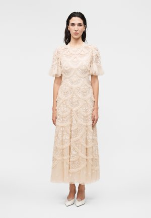 Donna che indossa un vestito midi in pizzo beige con maniche corte a sbuffo e tacchi bianchi a punta, in piedi davanti a uno sfondo chiaro e uniforme.