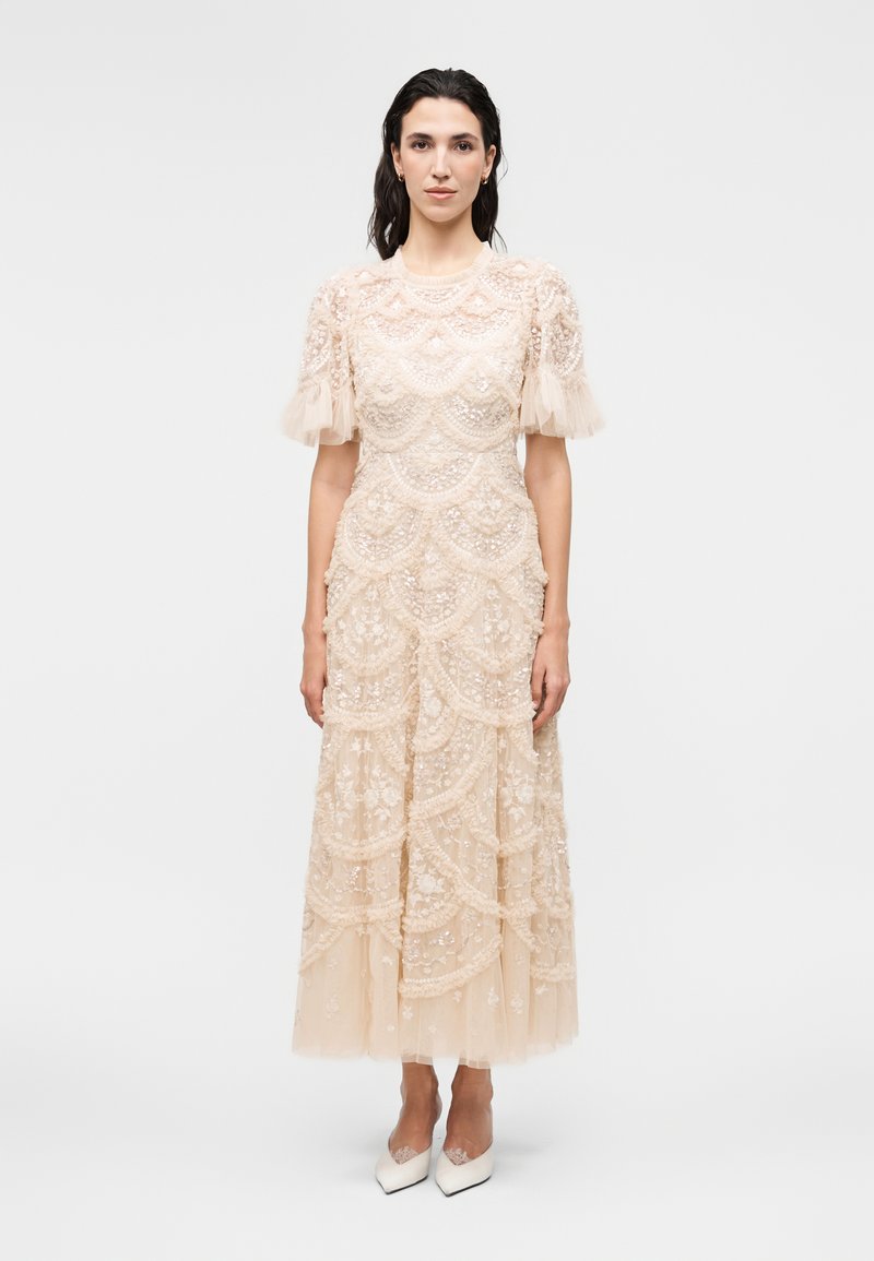 Γυναίκα που φοράει μπεζ δαντελένιο midi φόρεμα με κοντά φουσκωτά μανίκια και λευκά μυτερά τακούνια, στέκεται μπροστά σε απλό φωτεινό φόντο.