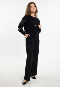 Cardigan nero con bottoni e maniche nere testurizzate caratterizzate da linee dorate. Abbinato a pantaloni neri e scarpe nere a punta.