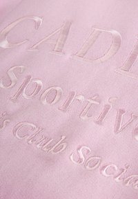 Różowa tkanina z wytłaczanym napisem "CADI Sportive Club Social". Tekstura jest gładka, z delikatnym połyskiem.