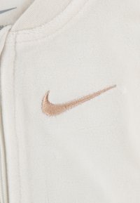 Fehér bolyhos ruházat, amelyen egy kiemelkedő bézs Nike pipa logó található. A textúra puhanak tűnik, zippzáras záródással a gallérnál.