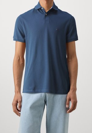 Mann trägt ein marineblaues kurzärmeliges Polohemd kombiniert mit hellblauen Jeans und steht vor einem schlichten hellen Hintergrund.