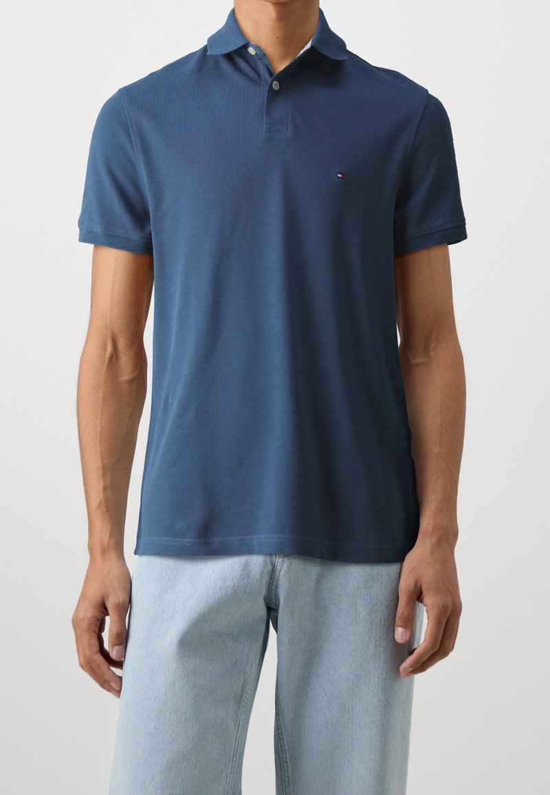 Homme portant un polo à manches courtes bleu marine associé à un jean denim bleu clair, debout devant un fond clair uni.