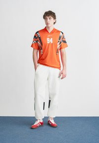 Orange atletisk tröja med svarta och vita grafiska detaljer, nummer 94 på framsidan. Vita byxor med svarta lappar, röda sneakers.