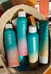 Vier bunte Coola-Sonnencremeprodukte in einem geflochtenen Korb auf einem gestreiften Stuhl im Freien.