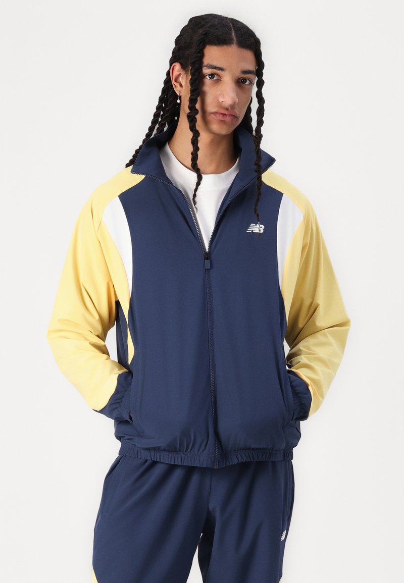 Giacca con zip blu navy e gialla con accenti bianchi, realizzata in materiale leggero. Presenta un colletto alto e una vita elasticizzata, con il logo del marchio.