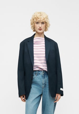 Person mit kurzen lockigen blonden Haaren, die ein marineblaues Sakko über einem weiß-rosa gestreiften Hemd und hellblaue, hoch taillierte Jeans trägt.