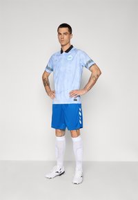 Maillot de football bleu clair avec un col noir et des manches à motifs, associé à un short bleu et des chaussettes blanches. Chaussures à crampons noires.