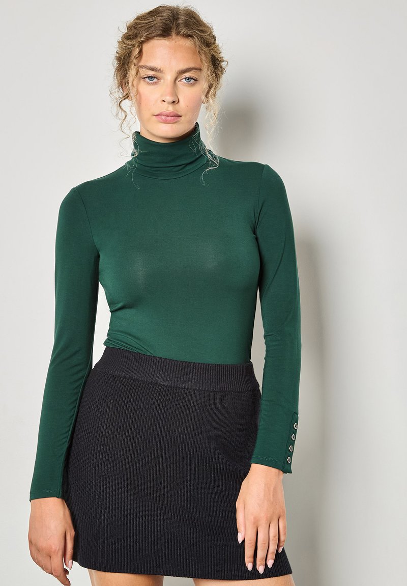 Maglia verde a collo alto e maniche lunghe abbinata a una mini gonna nera a coste. La gonna presenta un motivo testurizzato e dettagli con bottoni sui polsini della maglia.