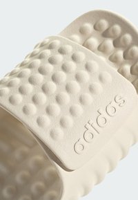 Adidas-sandaalit vaaleassa beige-kumissa, jossa on teksturoitu ja epätasaista yläremmi. Varustettu kohokuvioidulla brändilogolla. Muotoiltu pohja mukavuutta varten.