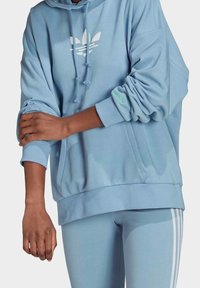 Ljusblå oversize-hoodie med vit logotyp, dragsko i huvan, muddade ärmar och grön detalj på ärmen; bärs med matchande leggings.