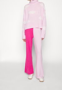 Rosa verkürzter Pullover mit weißen Wolkenmustern, kombiniert mit weit geschnittenen Hosen, die in zartem Rosa und leuchtendem Pink geteilt sind, getragen mit weißen Clogs.