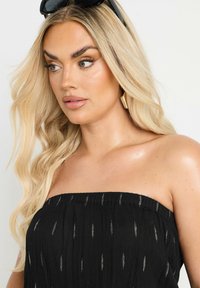 Svart strapless topp med vertikala silverdetaljer, i en texturerad, rynkad tyg. Modellen bär guldörhängen och svarta solglasögon.