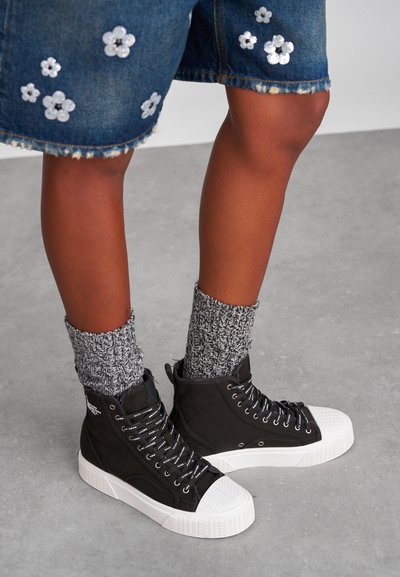 Nohy oblečené v jeanových šortkách po kolena s bílou květinovou výšivkou, šedými pletenými ponožkami a černými vysokými teniskami s bílou podrážkou a značkovými tkaničkami.