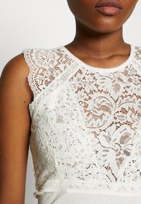 Närbild av en person som bär en ärmlös vit topp med intrikata blommönster av spets på bröstet och axlarna.