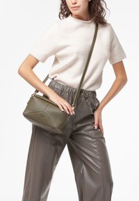 Olivenfarvet, struktureret læder crossbody taske med angular design, gyldent hardware og en justerbar rem, parret med let sweater og grå bukser.