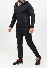 Ensemble de sport noir avec des poches zippées, tissu texturé, design ajusté et une capuche, associé à des baskets noires et orange.