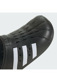 Scarpa nera con superficie testurizzata, dalla forma curvata, dotata di fori di ventilazione e tre strisce bianche sul lato. Realizzata in materiale sintetico resistente.