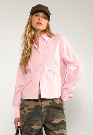 Woman wearing a light pink button-up shirt, camo pants, a dark brown cap, with long wavy blonde hair, standing against a plain background.