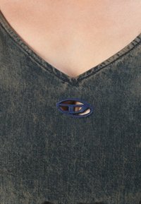 Modra denim bluza z V-izrezom, ki ima dekorativen morsko moder ovalni izrez na prsih. Nežen teksturiran videz.