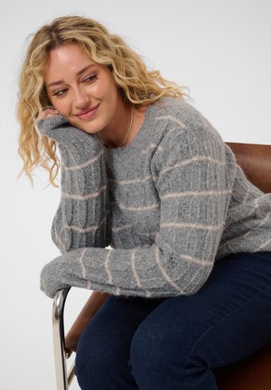 Femme aux cheveux blonds bouclés, portant un pull gris à motifs et un jean foncé, assise sur une chaise marron, appuyant sa tête sur sa main et souriant.