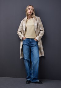 Beżowy wełniany płaszcz w kratę z klapami, jasnożółta koszula z długim rękawem i niebieskie dżinsy o szerokich nogawkach. Całość dopełniają czarne buty wsuwane.