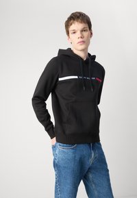 Svart hettegenser med justerbare snorer, frontlomme i kenguru-stil, og en horisontal stripe med merkevarenavnet i blå og røde detaljer.