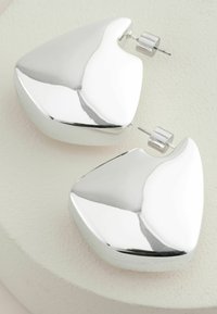 Cercei din metal cu nuanță argintie, cu o formă sculpturală, triunghiulară, având o suprafață satinat și sistem de prindere cu tortiță.