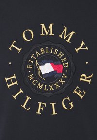 Černá tkanina s kulatou vyšívanou nášivkou s textem "TOMMY HILFIGER," zlatými akcenty a designem vlajky v červené, bílé a modré barvě.