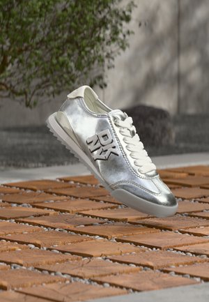 Srebrny sneaker z białymi sznurówkami i logo "DKNY" z boku, umieszczony nad drewnianą podłogą na zewnątrz, blisko rozmytej rośliny i ściany.