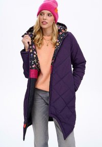 Fialová prošívaná bunda s květinovou podšívkou, kapucí a růžovými akcenty. Nosičena přes oranžový svetr a šedé džíny.