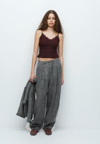 Burgundyröd texturerad crop top med vågiga kanter, tillsammans med lösa grå linnbyxor; modellen håller också en matchande jacka och bruna skor.