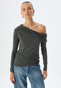 Dunkelgraues langärmliges, schulterfreies Oberteil mit drapiertem Ausschnitt und geraffter Textur, kombiniert mit hellblauen Denim-Jeans.