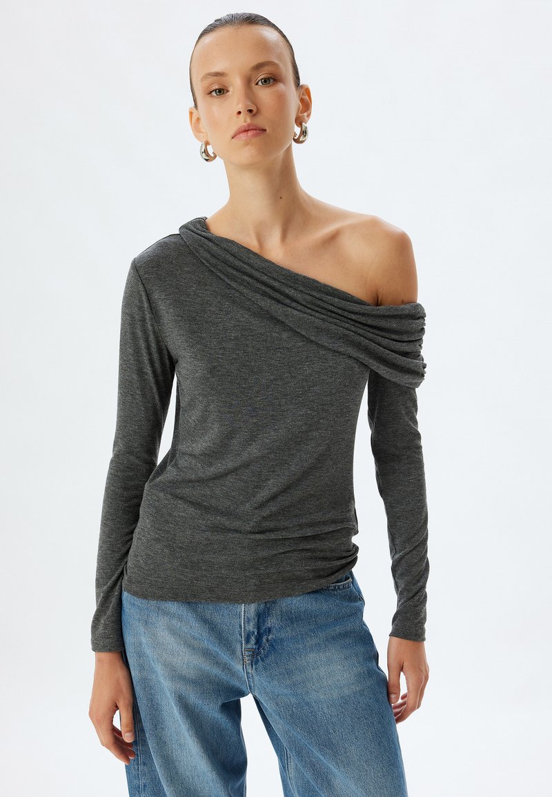 Dunkelgraues langärmliges, schulterfreies Oberteil mit drapiertem Ausschnitt und geraffter Textur, kombiniert mit hellblauen Denim-Jeans.