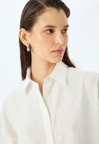 Camisa blanca con sutiles rayas verticales, cuello y cierre de botones. El modelo lleva pendientes de aro de plata suave. Textura suave, tejido ligero.