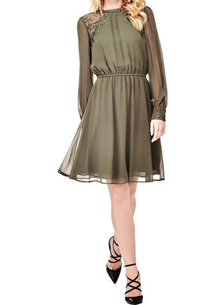 Femme portant une robe olive verte jusqu'aux genoux avec des manches longues transparentes, des détails en dentelle sur les épaules et les poignets, associée à des talons noirs à lanières.