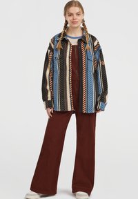 Camisa de franela a rayas multicolor con patrón de espiga y dos bolsillos frontales, combinada con pantalones de pana burdeos de pierna ancha.