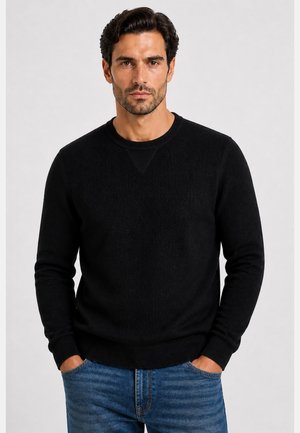 Mann mit dunklem Haar und hellem Bart, der einen schwarzen Rundhalspullover und blaue Jeans trägt, steht mit den Händen in den Taschen vor einem schlichten Hintergrund.