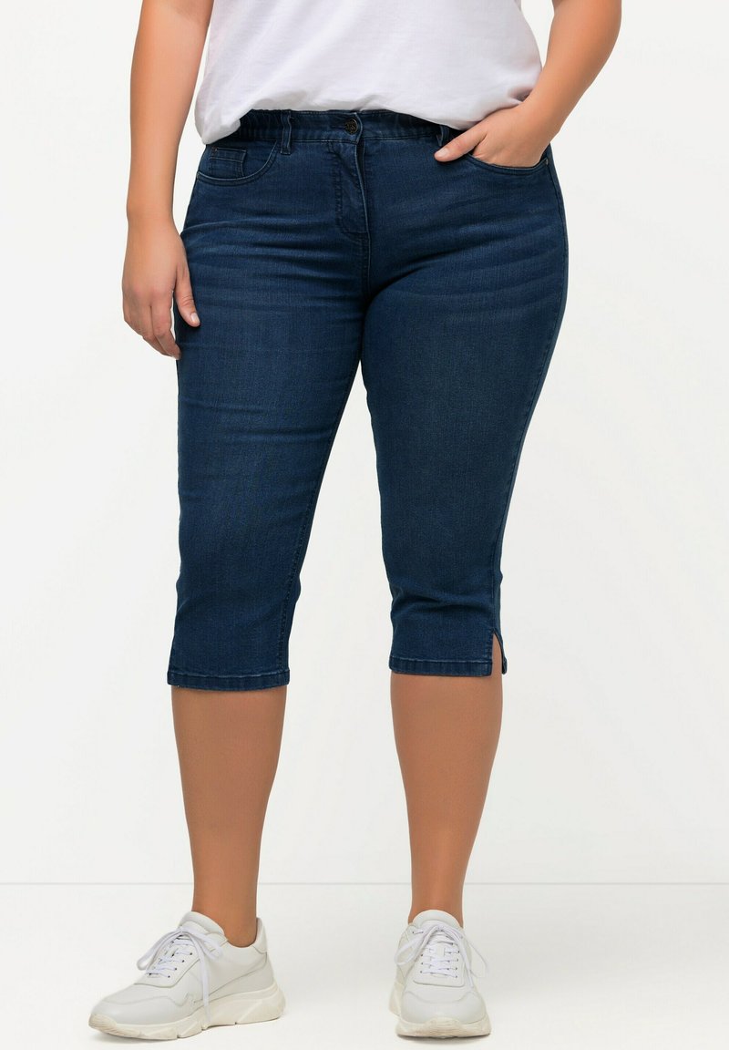 Pantalones capri de mezclilla azul oscuro con una textura suave, que presentan un cierre de botón y aberturas laterales en el dobladillo. Combinados con zapatillas blancas.