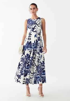 Frau trägt ein ärmelloses weißes Maxikleid mit großen dunkelblauen Blumenmustern, hält eine beige geflochtene Clutch und trägt weiße High Heels.