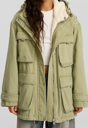 Personne portant un parka vert olive surdimensionné avec doublure en polaire, grandes poches avant, et un pull court blanc en dessous.