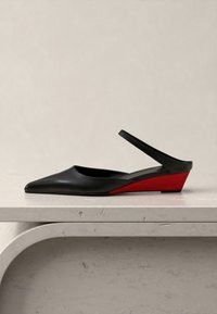 Μαύρο γυναικείο παπούτσι με μυτερή μύτη και λουρί στο πίσω μέρος, με χαμηλό κόκκινο φερόμενο τακούνι σε ελαφρώς λευκό μάρμαρο πάνω σε ουδέτερο φόντο.