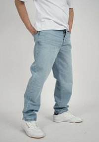 Person steht mit den Händen in den Taschen, trägt hellblaue Jeans, ein weißes T-Shirt und weiße Reebok-Sneaker vor einem einfarbigen Hintergrund.