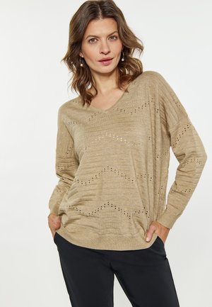 Beige kötött pulóver V nyakkal, textúrált mintázattal és kisebb kivágásokkal. Hosszú ujjú és laza szabású. Sötét nadrággal párosítva.