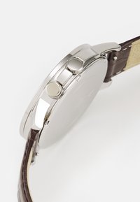Montre en acier inoxydable avec un bracelet en cuir marron. Elle présente deux boutons sur le côté et une finition polie, mettant en valeur un design épuré.