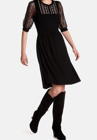 Robe noire avec des manches bouffantes en dentelle translucide à motifs, un corsage ajusté et une jupe évasée arrivant au genou ; complétée par des bottes noires mi-mollet.