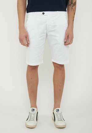 Uomo con pantaloncini bianchi fino al ginocchio, camicia nera e scarpe da ginnastica bianche con strisce nere, in piedi davanti a uno sfondo chiaro e uniforme.