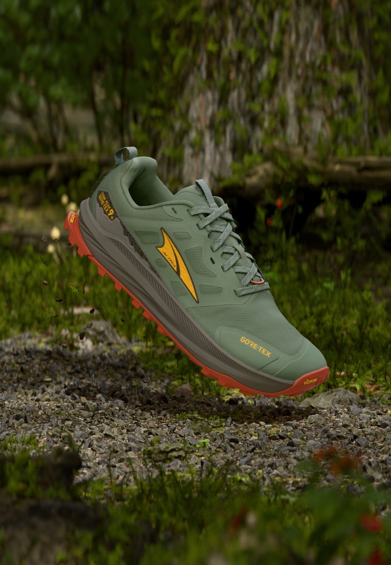 Chaussure de trail Gore-Tex verte et orange flottant au-dessus de gravier et d'herbe dans un environnement forestier.