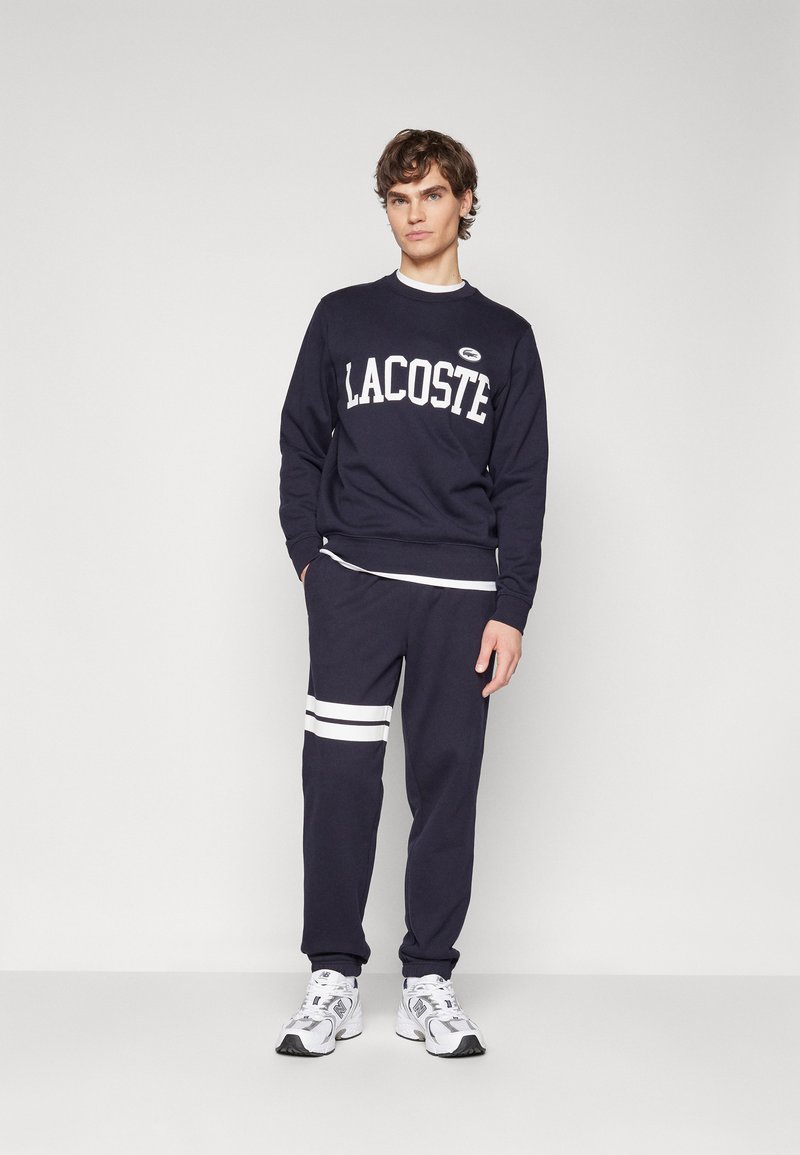 Sweatshirt bleu marine avec logo "LACOSTE" blanc et pantalon assorti présentant des rayures blanches au niveau du genou. Le tissu semble doux et décontracté.